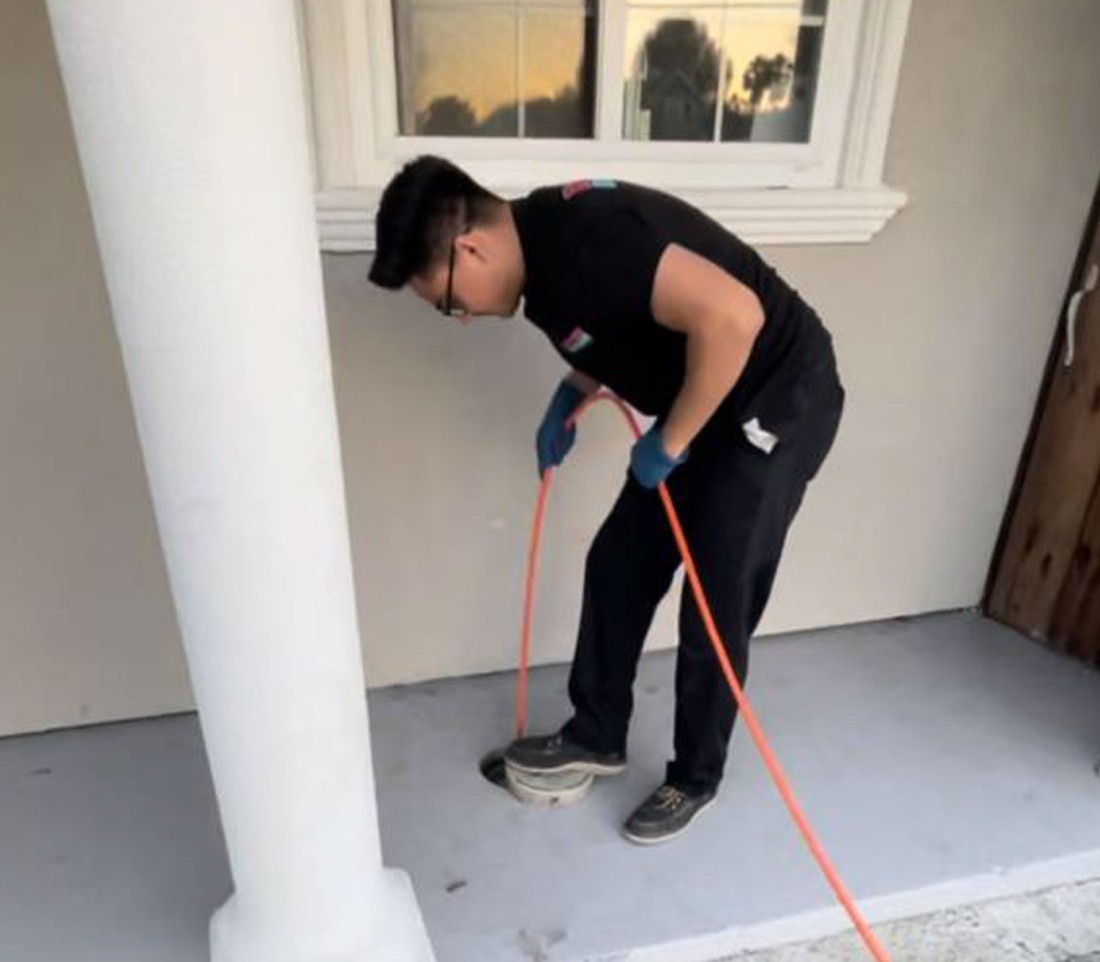 Person cleaning a porch floor with an orange hose, next to a white column and under a window.