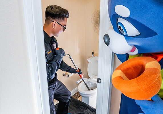 Plumber fixing toilet with mascot character watching in a bathroom.