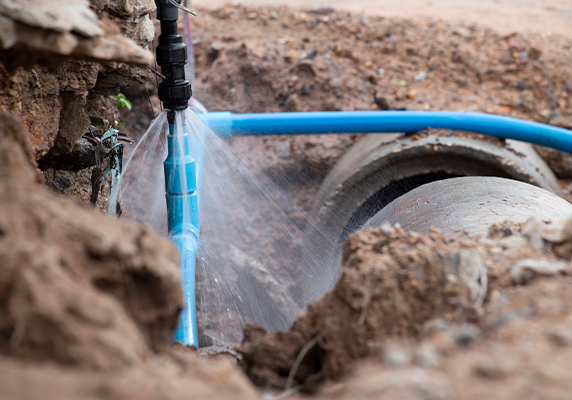 Leaking blue water pipe in a muddy trench indicates need for repair, highlighting infrastructure issues.