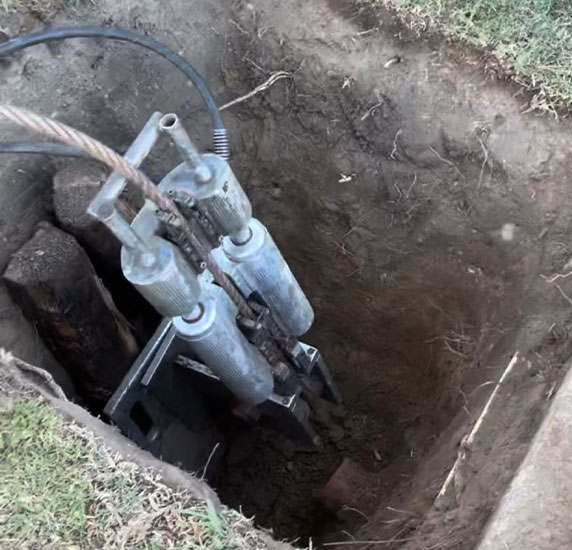 Hydraulic log splitter vertically positioned in a dug hole, showing the machine's mechanism and surrounding soil.