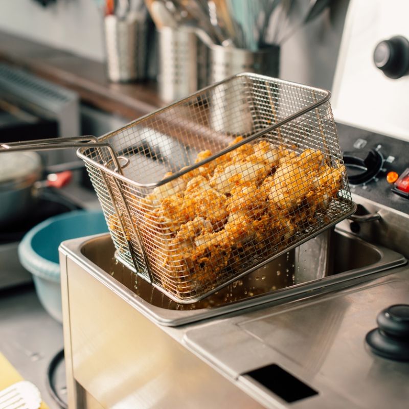 Fried chicken in a fryer basket being lifted from hot oil in a commercial kitchen setting.