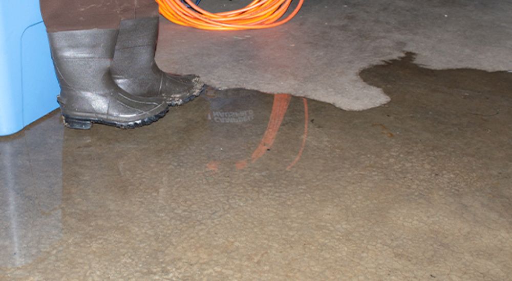 Person in boots standing near water puddle on garage floor, holding an orange extension cord.