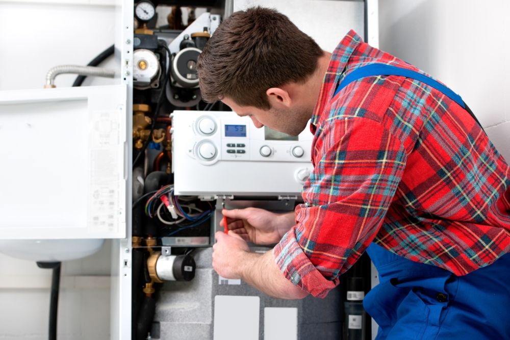 Technician repairing a home boiler system, adjusting controls, wearing plaid shirt and overalls in a utility room.