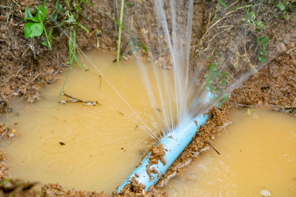 Leaking blue pipe in muddy ground, water spraying out forcefully, causing erosion and flooding the area.