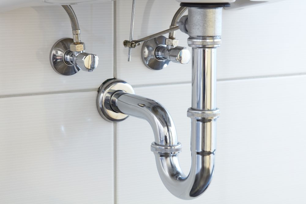 Shiny chrome sink plumbing and water shut-off valves on a tiled bathroom wall.