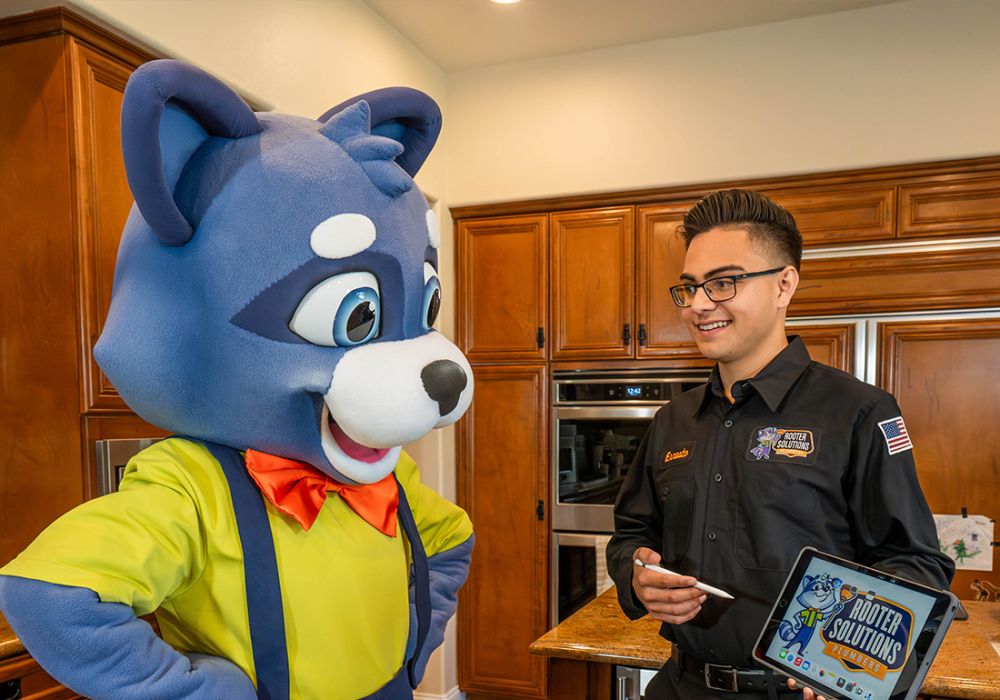 Mascot and technician discuss plumbing solutions in kitchen, featuring tablet with Rooter Solutions logo.