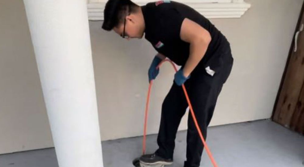 Person cleaning a porch floor with an orange hose, next to a white column and under a window.