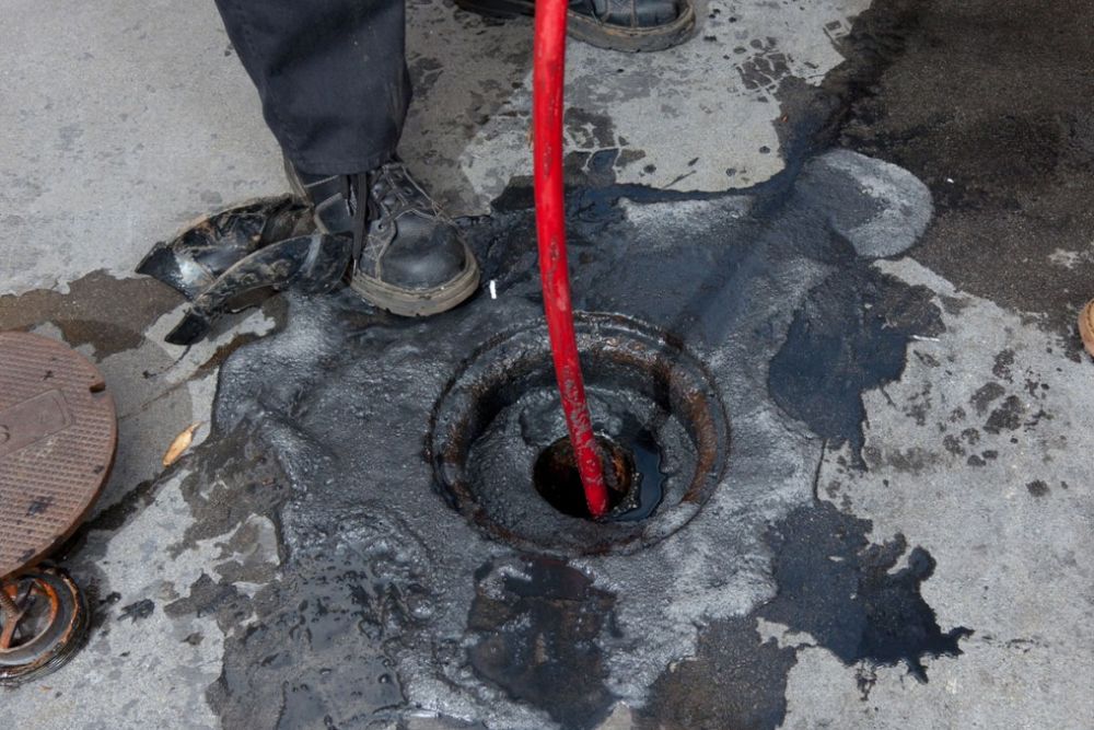 Worker cleaning blocked drain with red hose, surrounded by spilled black liquid on concrete.