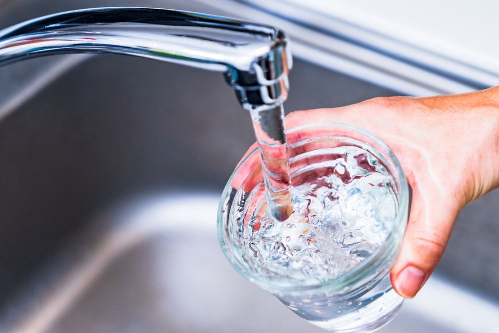 Hand holding glass under running faucet, filling with fresh tap water in a kitchen sink.