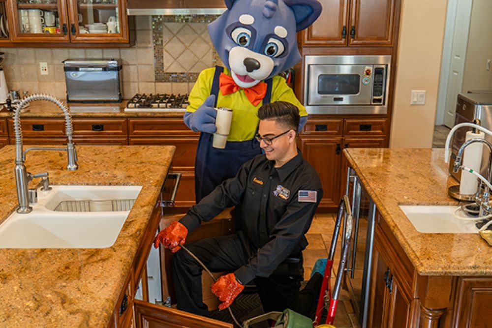 Plumber fixing kitchen sink as mascot watches with a drink, showcasing fun and efficient service.