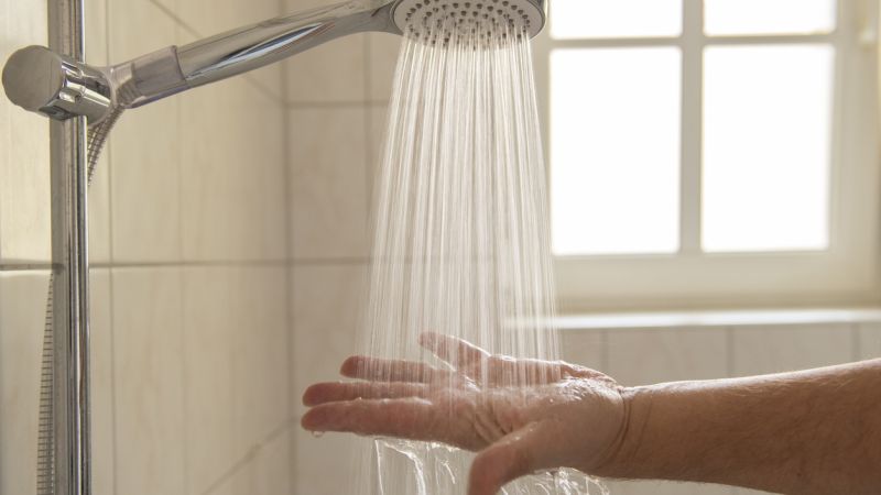 Hand testing water from showerhead in bright bathroom with tiled walls and window in background.