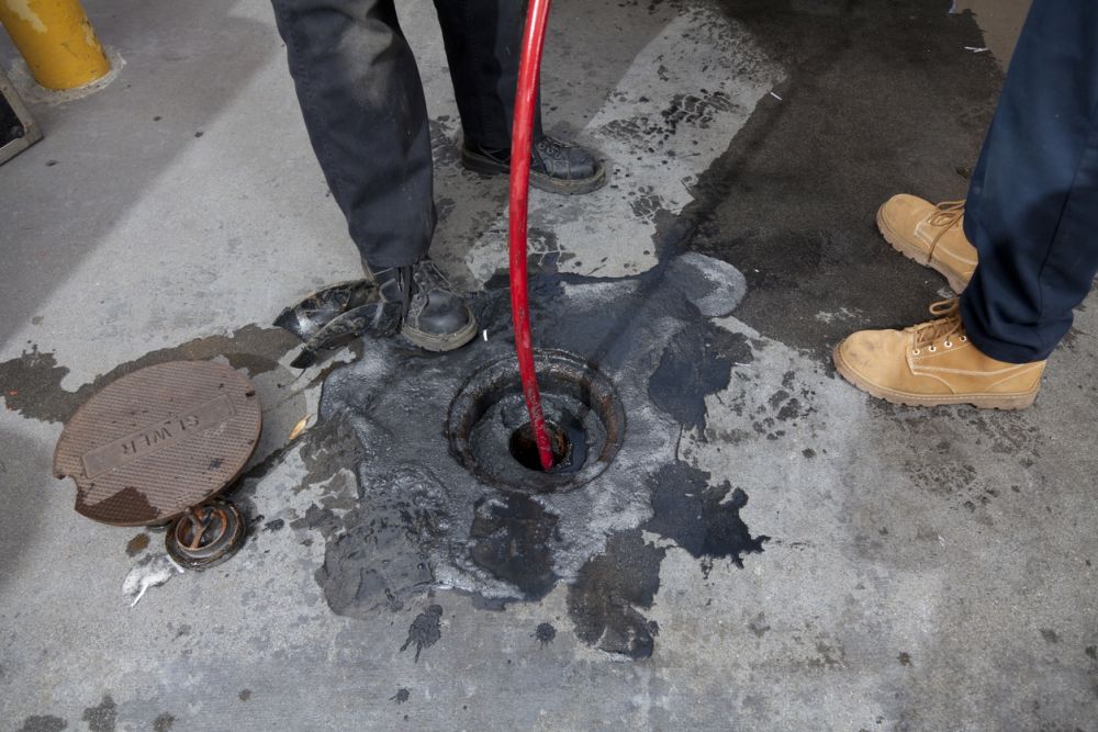Workers using red hose for sewer maintenance; shoes and open manhole visible on concrete surface.
