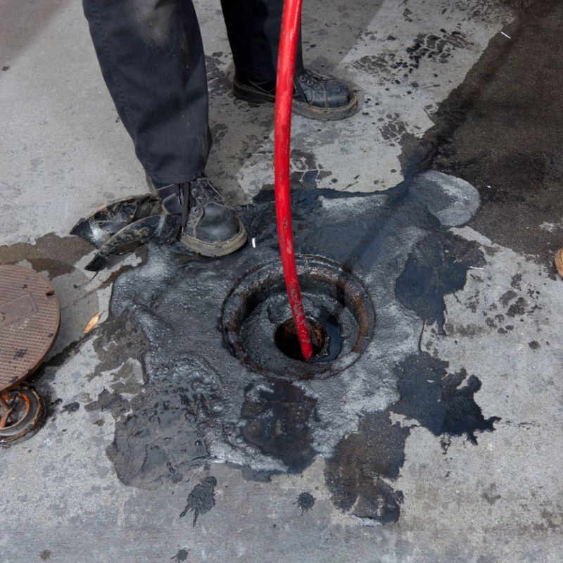 Worker cleaning blocked drain with red hose, surrounded by spilled black liquid on concrete.