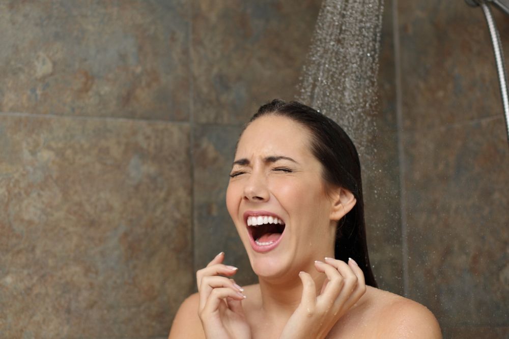Woman in shower reacting to cold water, expressing surprise, under a flowing showerhead on a tiled background.