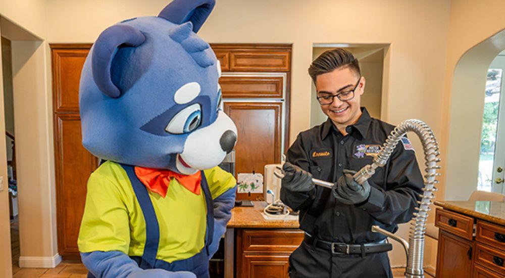 Cartoon mascot and technician fixing kitchen faucet in a bright home setting.