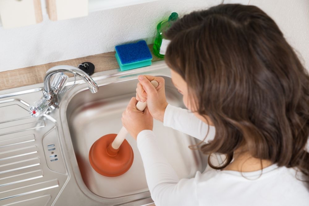 Woman using plunger in kitchen sink for cleaning blocked drain, surrounded by sponge and detergent bottle.