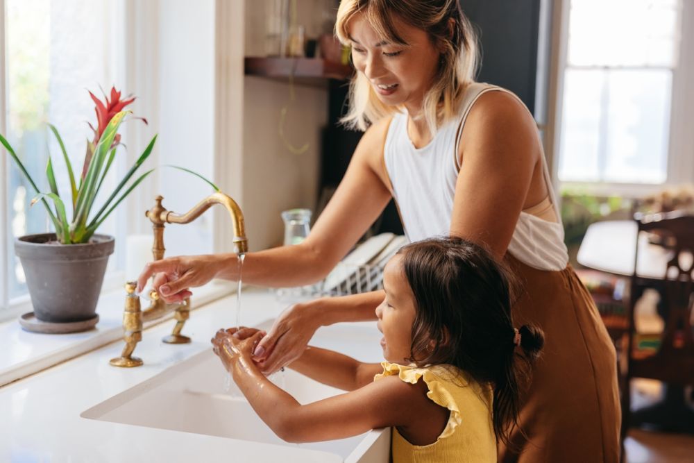 Mother and daughter washing hands together at the kitchen sink, reinforcing hygiene and family bonding.