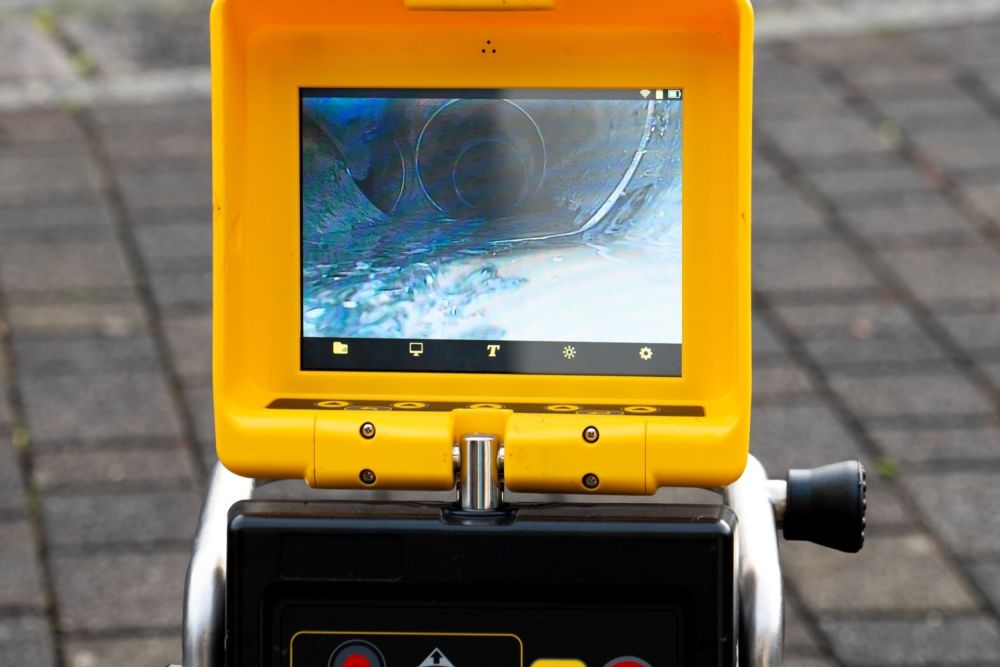 Plumbing inspection camera displaying sewer pipe view on a yellow screen, positioned outdoors on a paved surface.