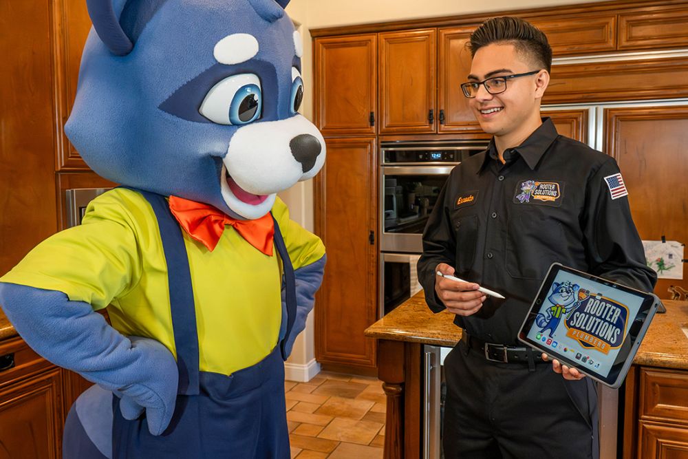 Mascot and technician discuss plumbing solutions in kitchen, featuring tablet with Rooter Solutions logo.