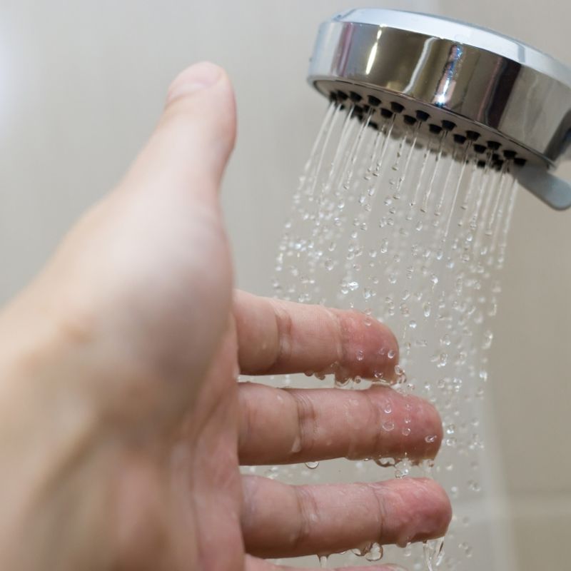 Hand testing water temperature from shower head, silver bathroom fixture, close-up of water droplets.