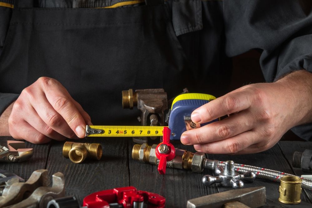 Plumber measuring pipe fittings with tape measure on a workbench surrounded by plumbing tools.