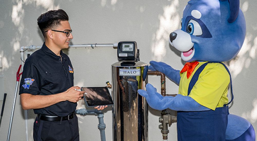 Technician and raccoon mascot discussing water softener installation outside.
