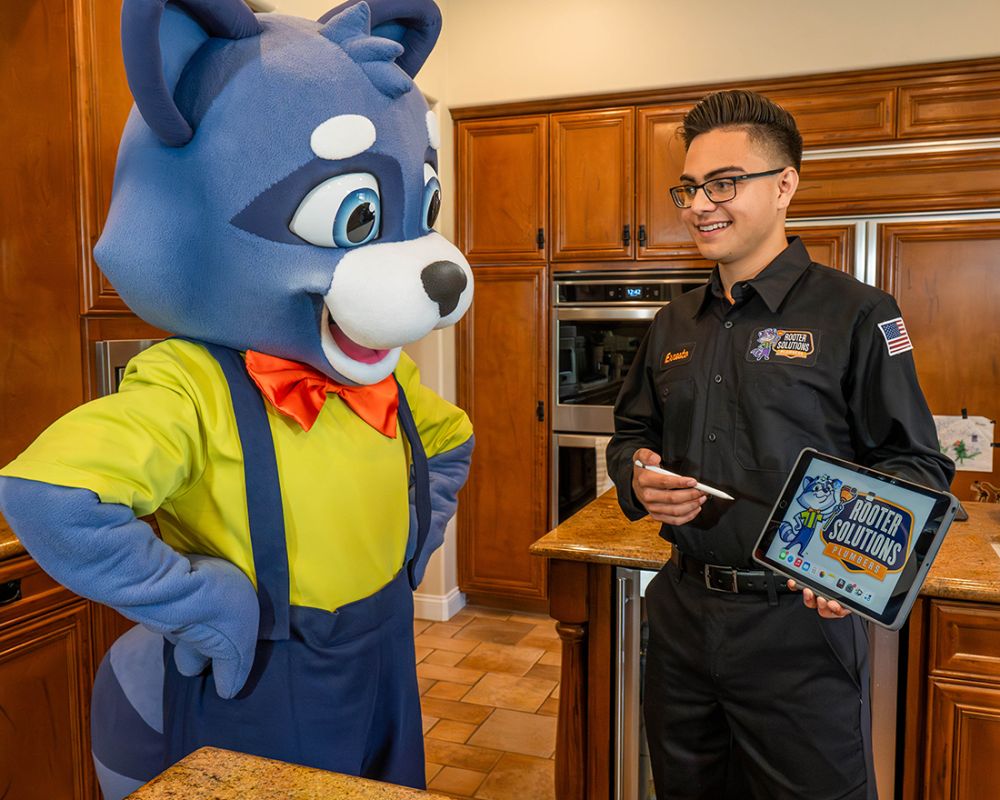 Mascot and technician discuss plumbing solutions in kitchen, featuring tablet with Rooter Solutions logo.