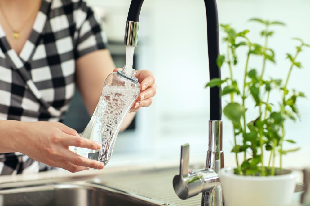 Woman filling reusable bottle with tap water in a kitchen next to fresh herbs. Eco-friendly lifestyle concept.
