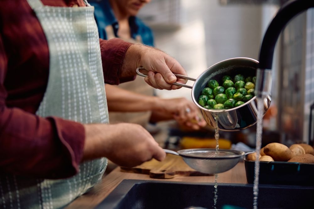 Person draining Brussels sprouts in a kitchen, with apron and a sink, preparing a meal.