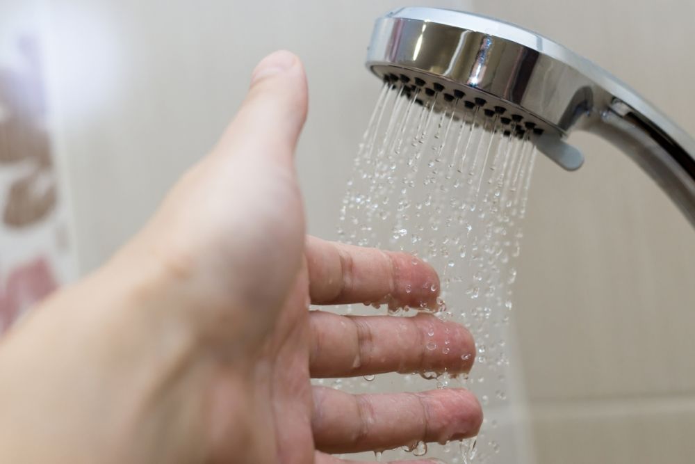 Hand testing water temperature from shower head, silver bathroom fixture, close-up of water droplets.