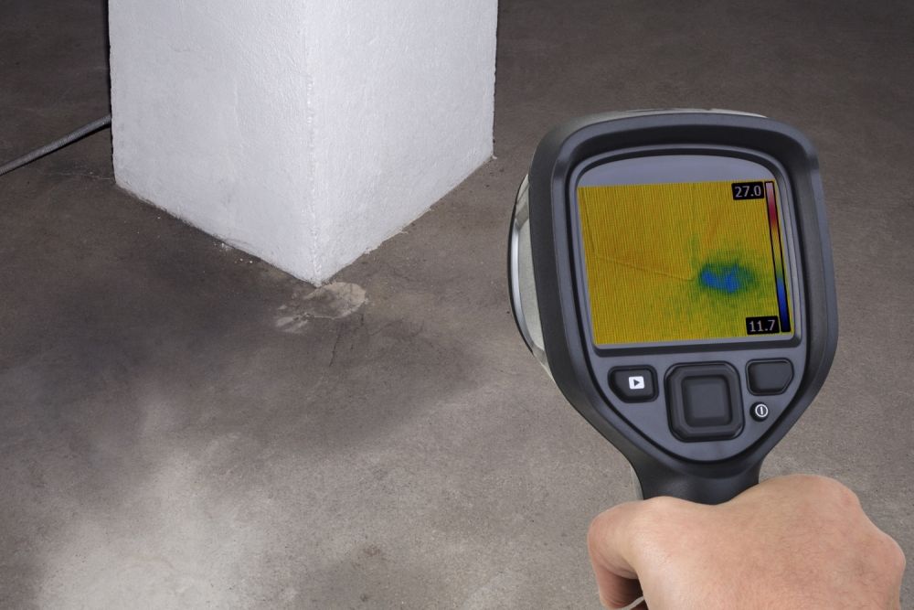 Thermal imaging camera detecting moisture near a concrete column, highlighting temperature differences on a screen.
