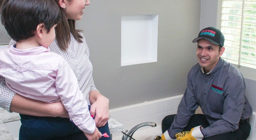 Plumber servicing a bathroom sink while a mother and child look on, illustrating friendly home repair service.