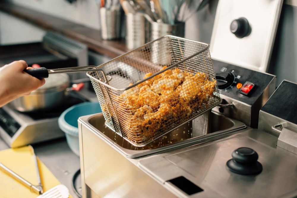 Fried chicken in a fryer basket being lifted from hot oil in a commercial kitchen setting.