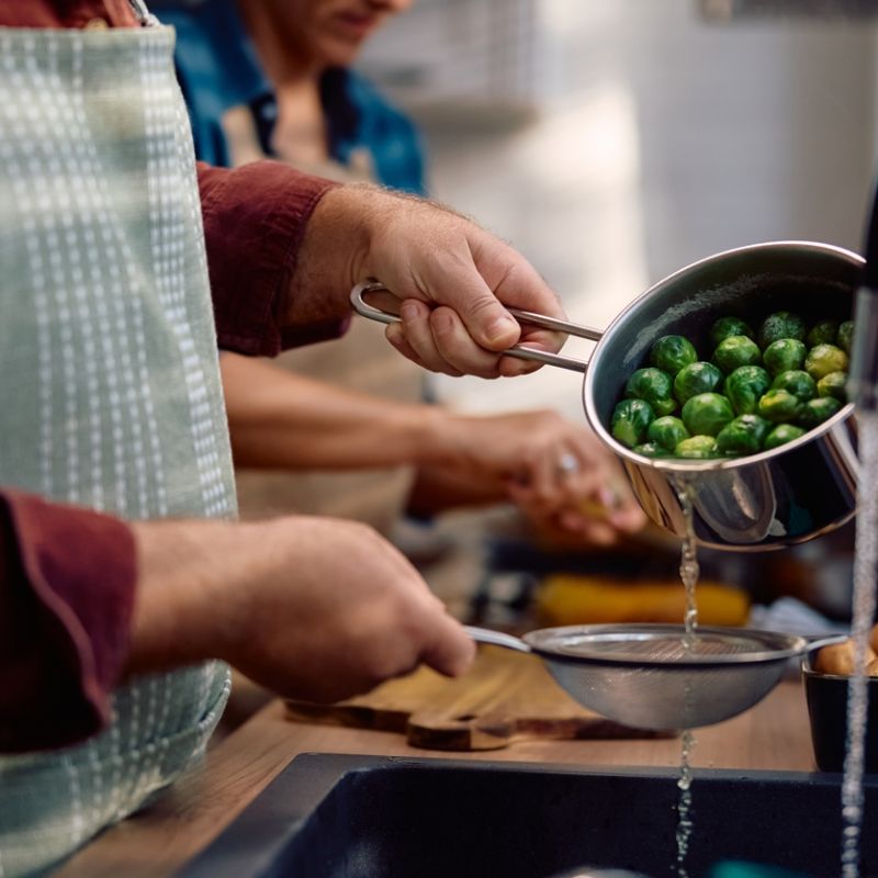 Person draining Brussels sprouts in a kitchen, with apron and a sink, preparing a meal.