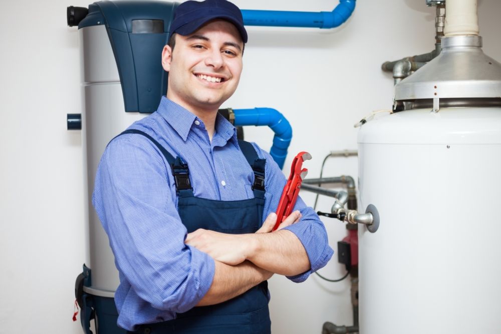 Smiling plumber holding wrench near water heater and pipes in boiler room.