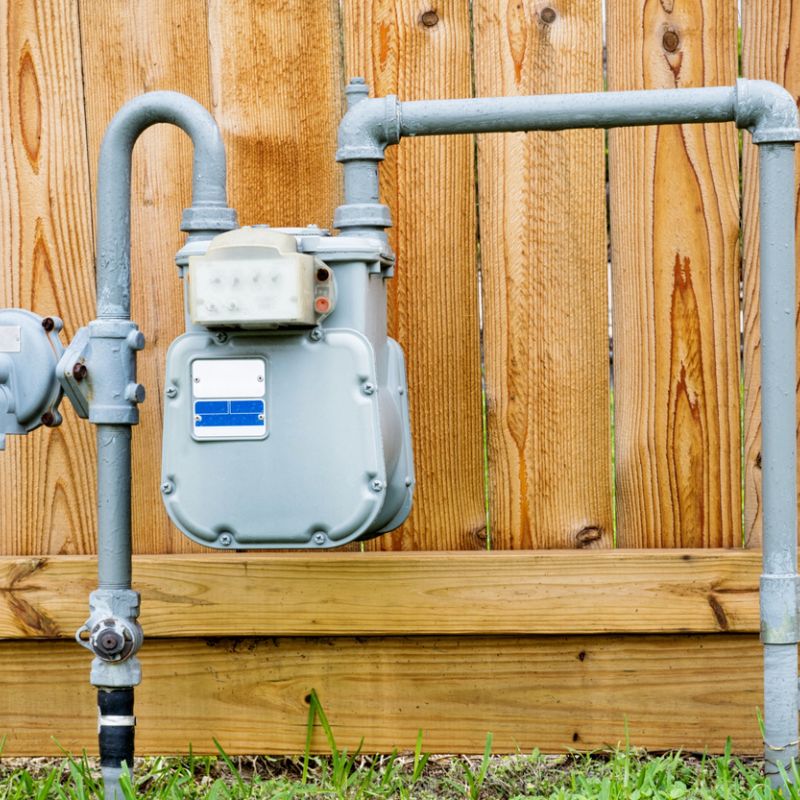 Outdoor residential gas meter with pipes against a wooden fence.