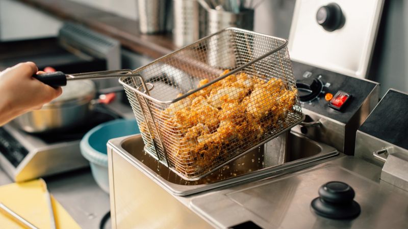 Fried chicken in a fryer basket being lifted from hot oil in a commercial kitchen setting.