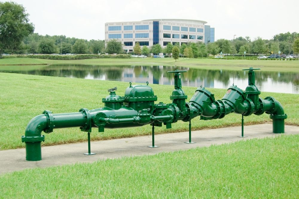 Green industrial pipes in a grassy area near a pond with a modern office building in the background.