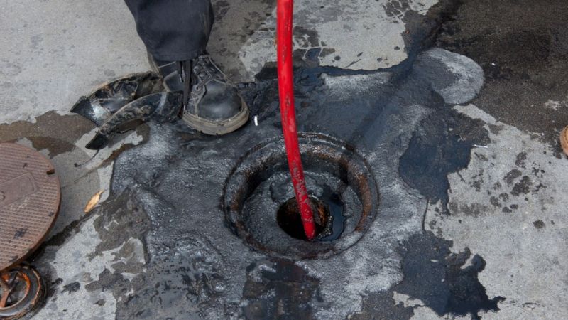 Worker cleaning blocked drain with red hose, surrounded by spilled black liquid on concrete.