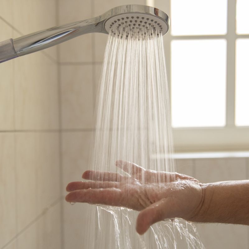 Hand testing water from showerhead in bright bathroom with tiled walls and window in background.