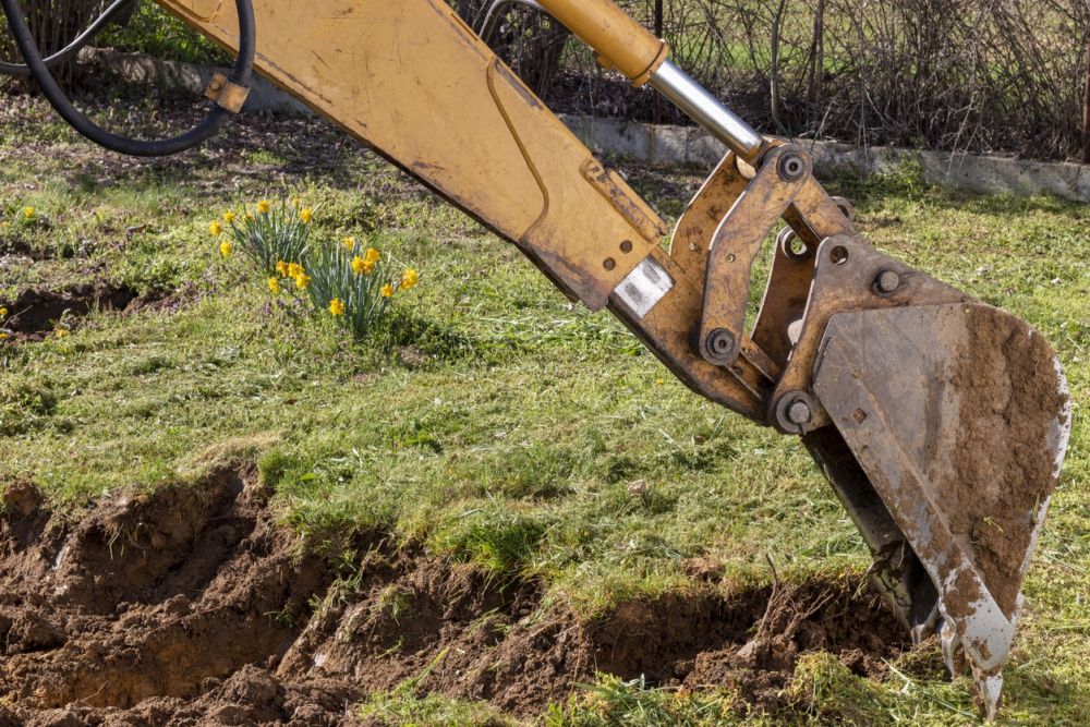 Excavator digging soil in a garden with blooming yellow flowers, spring construction site.