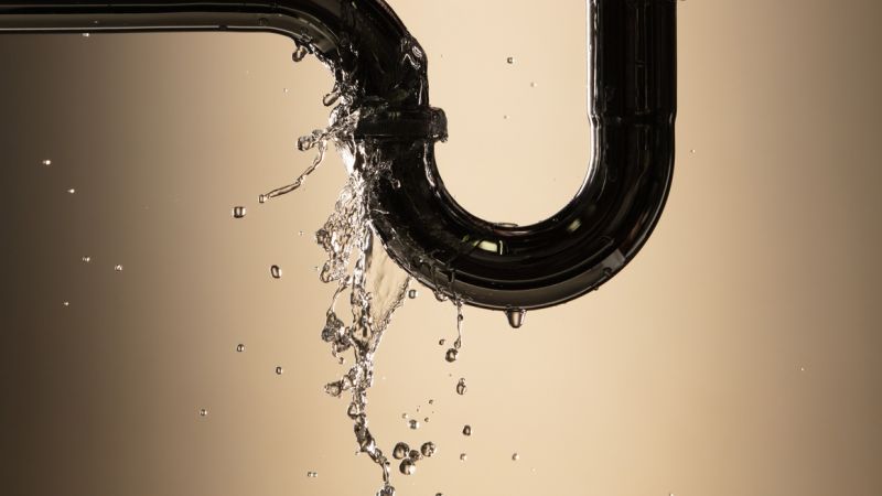 Leaking pipe with water flow, beige background, highlighting plumbing issues and repair needs.