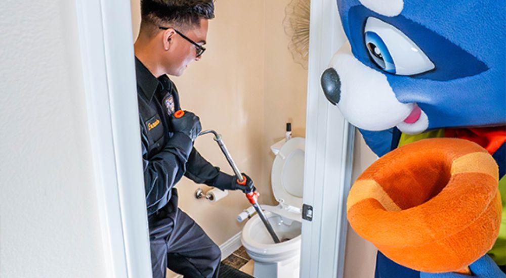 Plumber fixing toilet with mascot character watching in a bathroom.