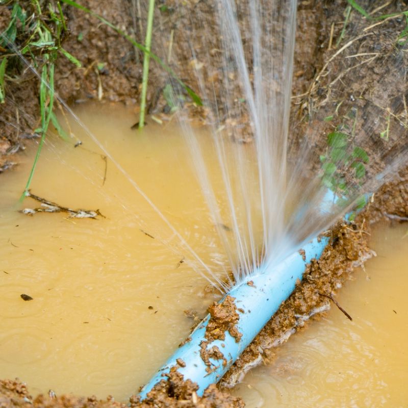 Leaking blue pipe in muddy ground, water spraying out forcefully, causing erosion and flooding the area.