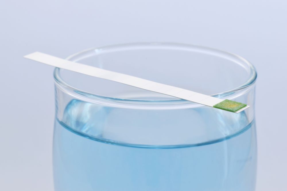 pH test strip on glass of blue liquid, indicating water acidity or alkalinity.