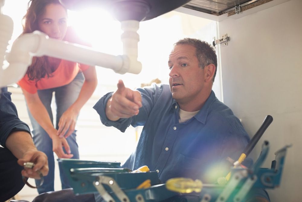 Plumber repairing sink pipes under kitchen cabinet while woman looks on.