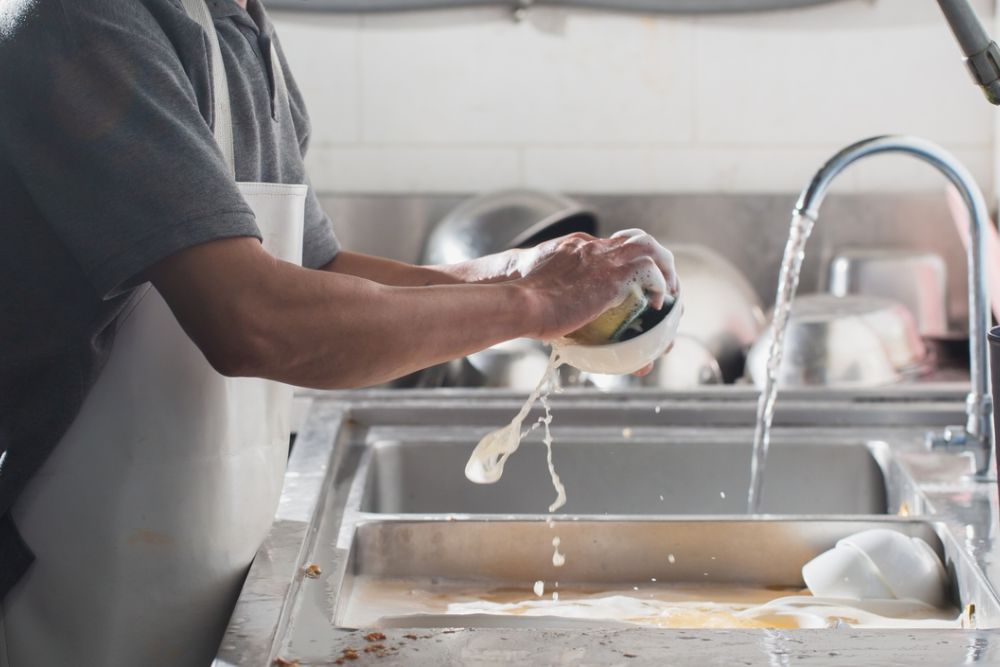 Person washing dishes at a sink, rinsing soapy cups under running water. Clean kitchen routine.