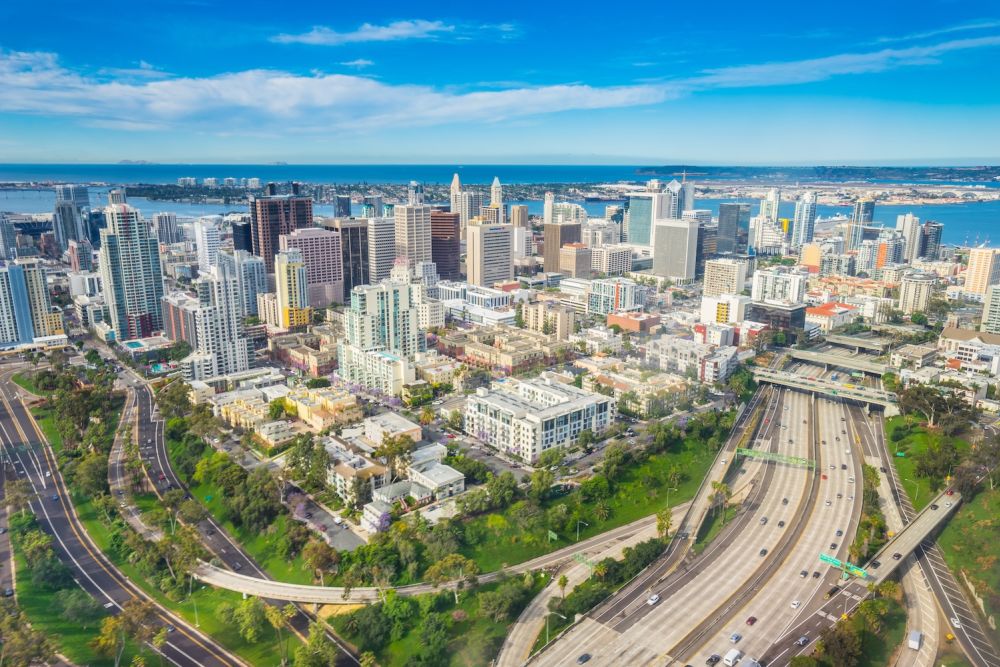 Aerial view of San Diego skyline with skyscrapers, highway, and coastline under a clear blue sky.