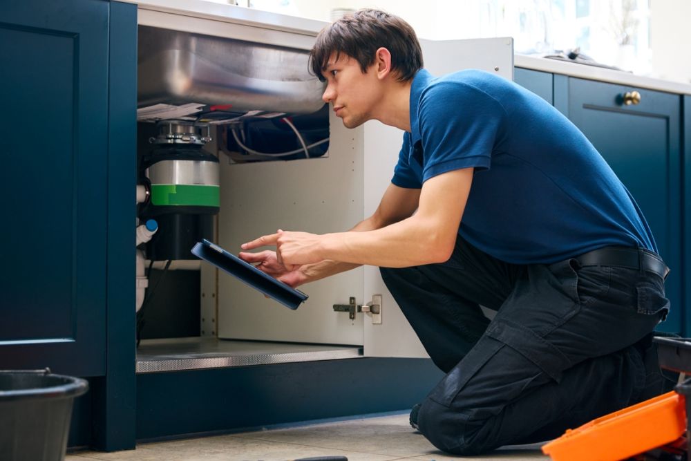Plumber troubleshooting under-sink issue using tablet, surrounded by tools in a modern kitchen setting.