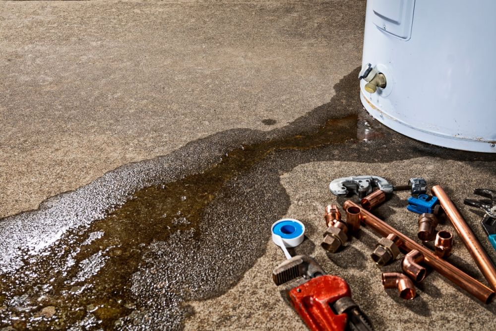 Leaking water heater with plumbing tools and repair parts on concrete floor.